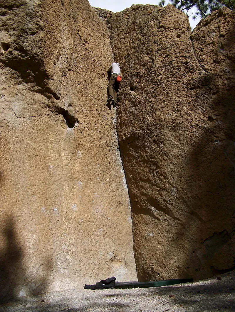 A climber high in a crack between two tall golden boulders, with a crash pad on the ground far below
