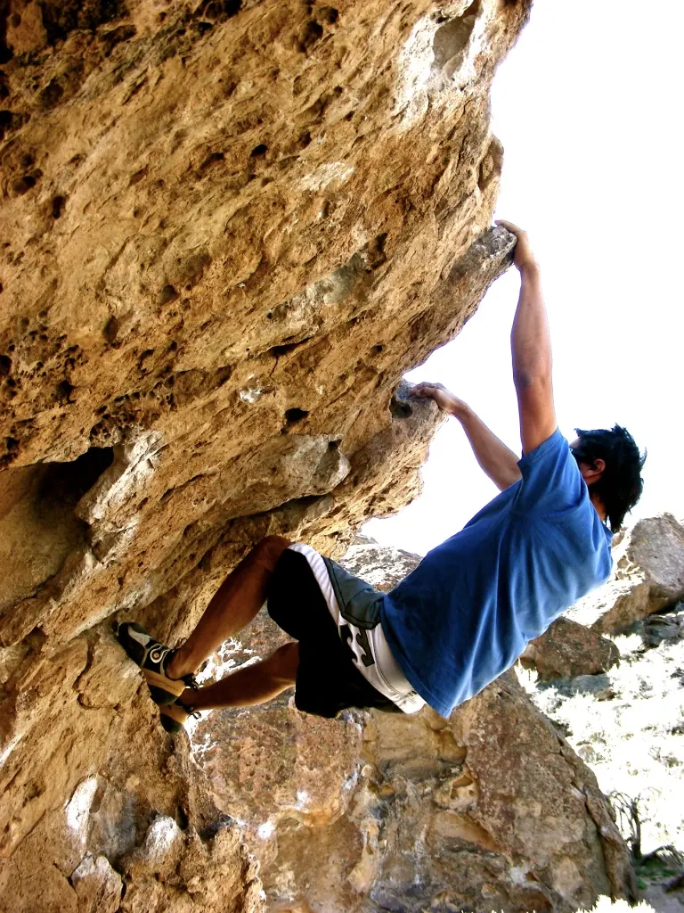 A boulderer in a blue shirt reaching up the underside of a steep, pocketed volcanic overhang