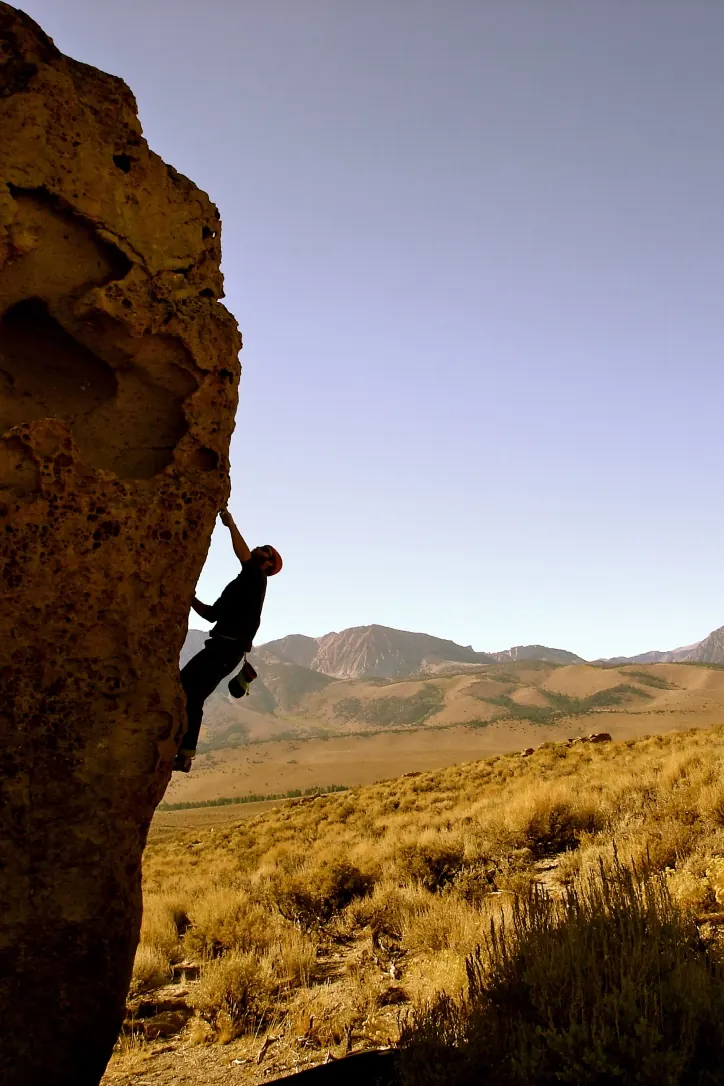 A climber silhouetted on the edge of a tall pocketed boulder in the sagebrush, with the Mono Basin hills and Sierra beyond