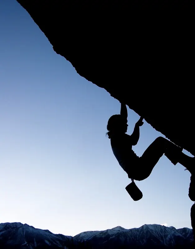 A climber silhouetted under a steep overhang at dusk, with snowy Sierra peaks on the horizon