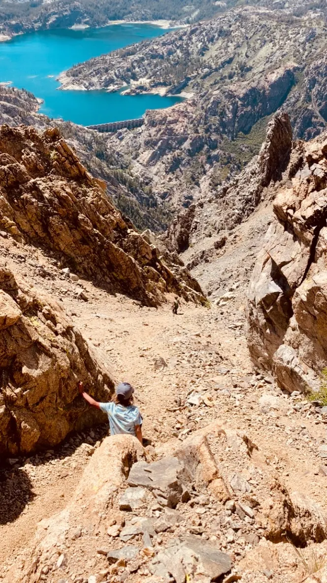 A child sliding down a steep gravel scree chute on the back side of Carson Peak, with a turquoise reservoir far below