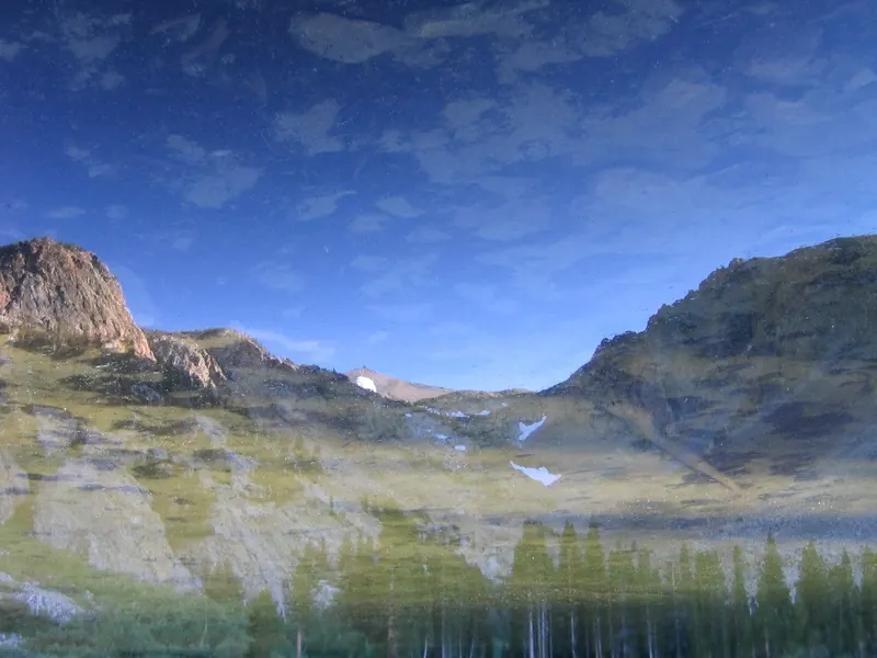 The cirque above Fern Lake reflected upside down in the still water: granite, snow patches and pines under a blue sky
