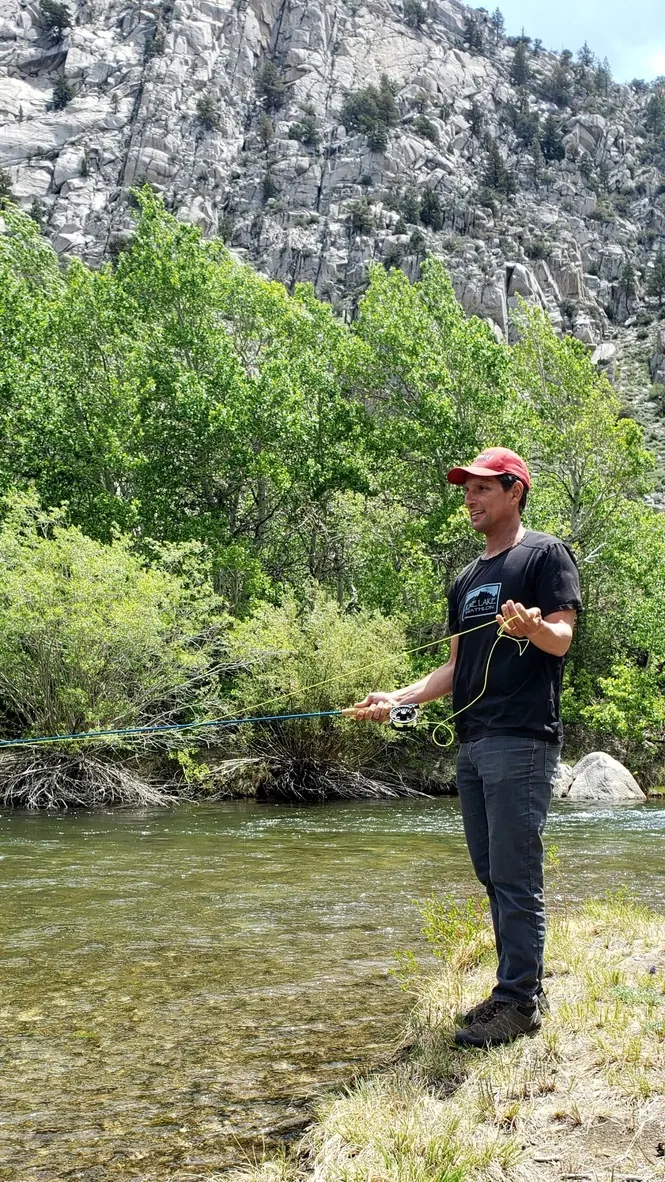 An angler in a red cap stripping in line beside a clear creek below granite cliffs and spring aspens