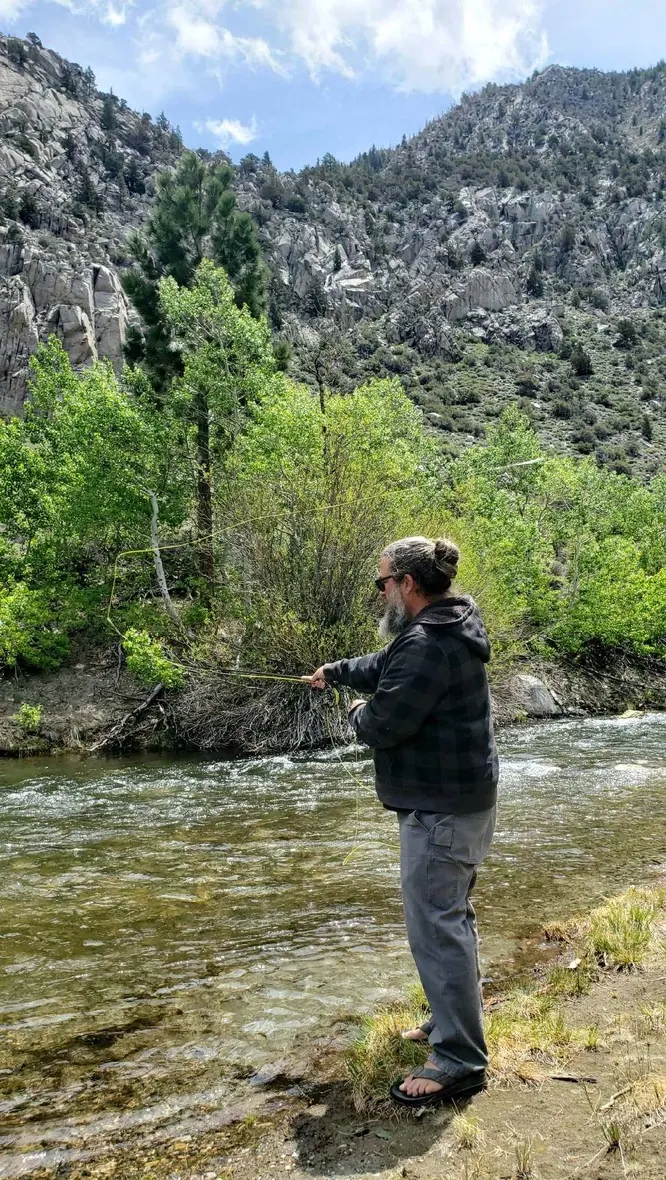 A bearded angler in a flannel hoodie casting a fly rod on a clear freestone creek below granite cliffs and aspens