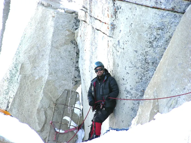 A climber in a black down jacket and helmet grinning at a belay against a granite wall, ropes running through the snow