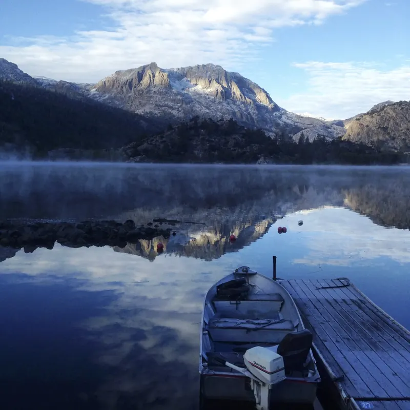A small aluminum rental boat tied to a wooden dock on a mirror-still lake, with morning mist and a snow-dusted granite peak reflected