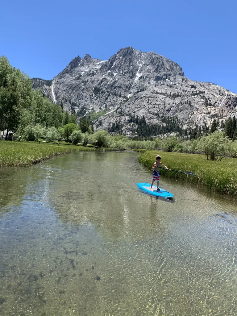 A young boy standing on a small blue paddleboard on clear Rush Creek, under Carson Peak