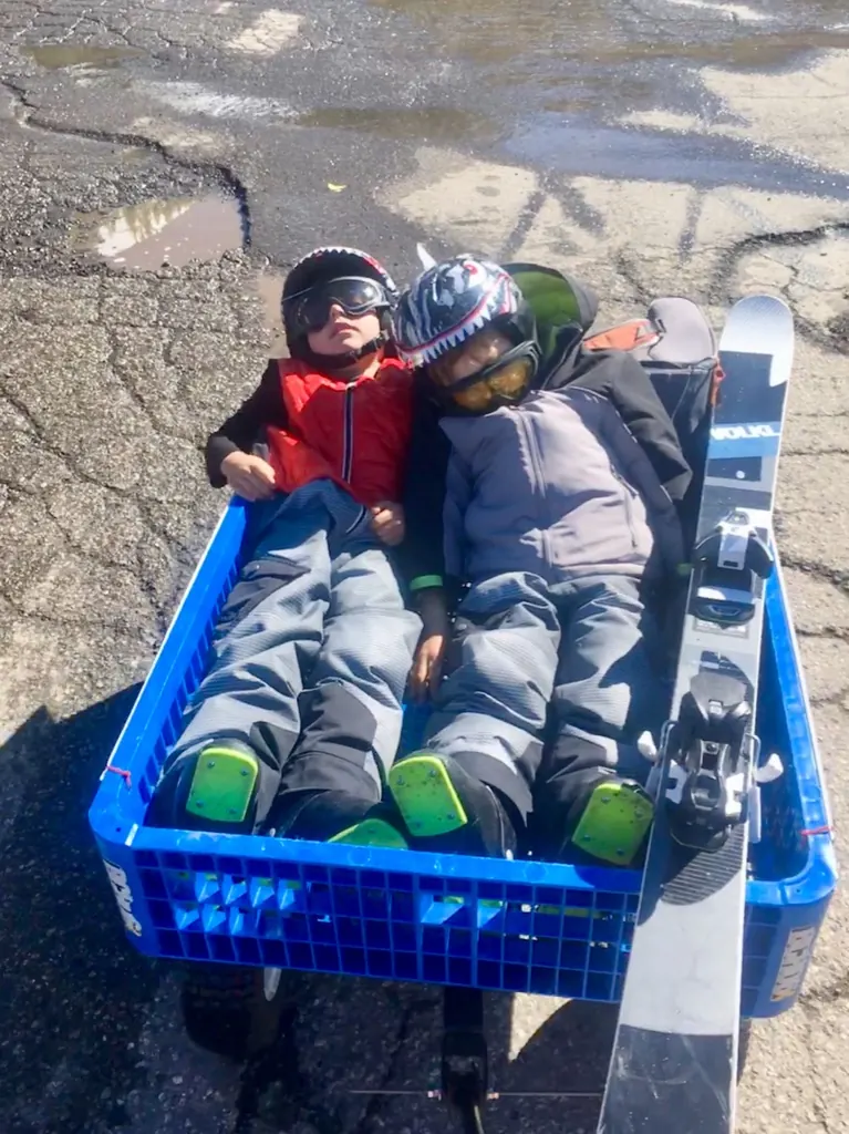 Two young skiers in helmets and goggles riding home in a gear cart beside a pair of skis