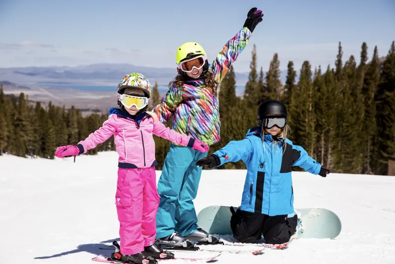Three kids grinning on a sunny June Mountain run, two on skis and one kneeling with a snowboard, with Mono Lake far below