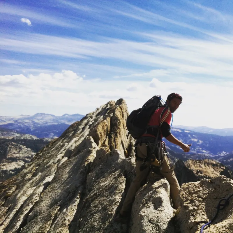 A climber in a harness and pack on the knife-edge granite of Matthes Crest, with the Yosemite high country behind