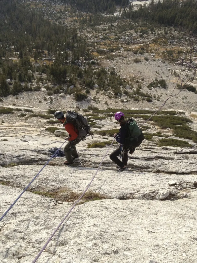 Two climbers with packs and helmets rappelling down a granite slab on Matthes Crest, with forest and meadow far below