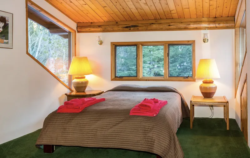 The River Room's queen bed with two lamps glowing, under a pine ceiling, with a window into the pines and an angled window onto the aspens