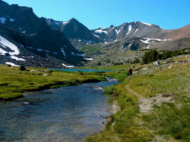 A hiker on a trail beside a clear creek, with an alpine lake and snow-streaked peaks ahead