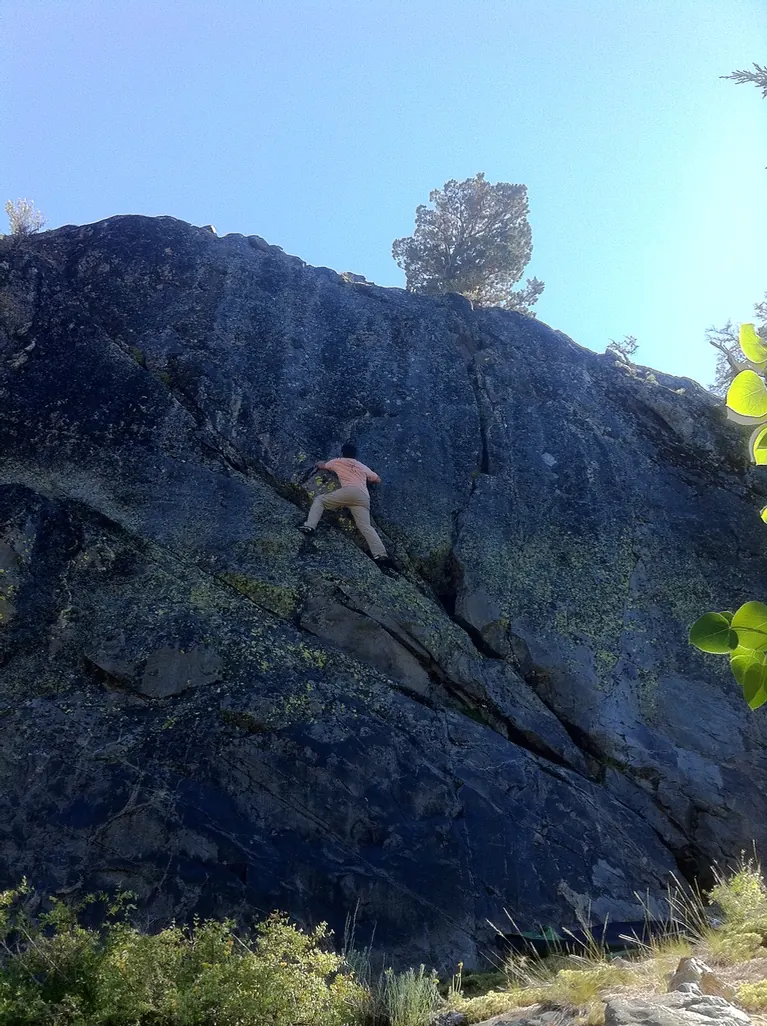 A climber in an orange shirt working up a huge dark lichen-streaked slab under a blue sky, an aspen leaf in the corner